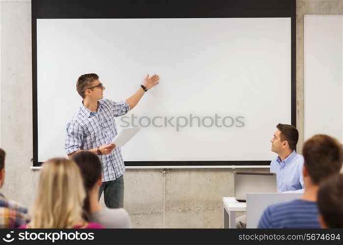 education, high school, technology and people concept - smiling student boy in glasses with notepad, laptop computer standing in front of students and teacher in classroom