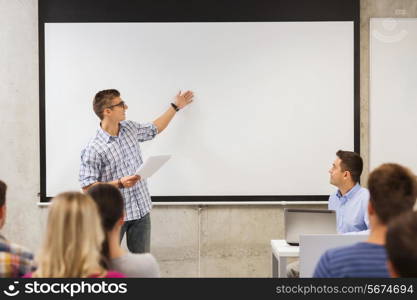 education, high school, technology and people concept - smiling student boy in glasses with notepad, laptop computer standing in front of students and teacher in classroom