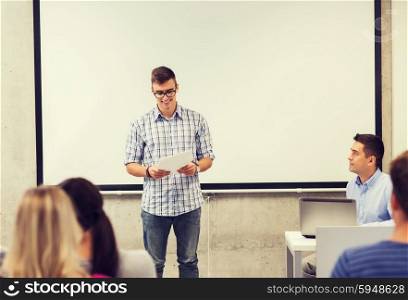 education, high school, technology and people concept - smiling student boy in glasses with notepad, laptop computer standing in front of students and teacher in classroom