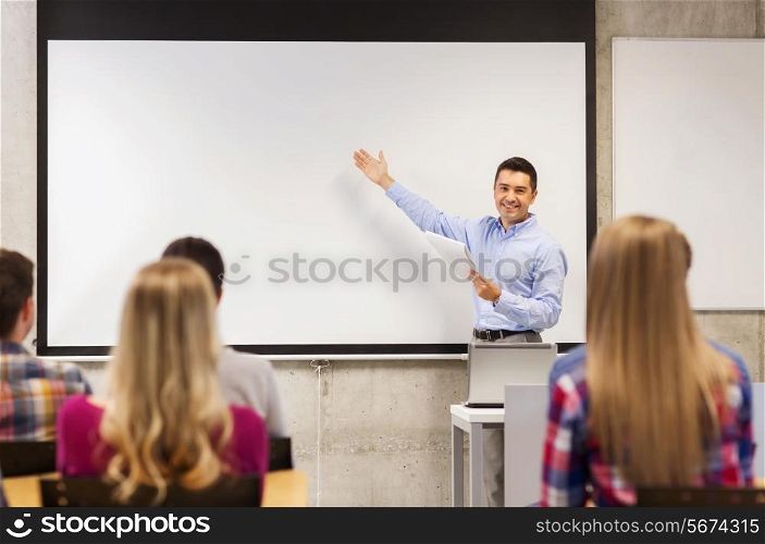education, high school, technology and people concept - smiling teacher with notepad, laptop computer standing in front of students and showing something on white board in classroom