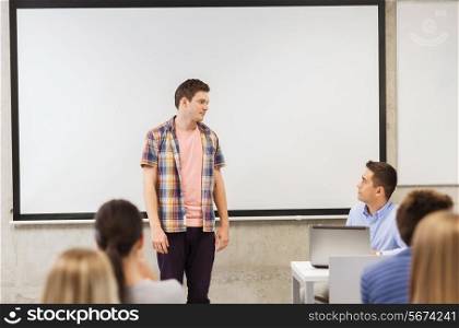 education, high school, technology and people concept - student boy standing in front of students and teacher with laptop computer in classroom