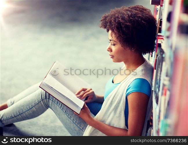 education, high school, university, learning and people concept - african american student girl reading book sitting on floor at library. african student girl reading book at library