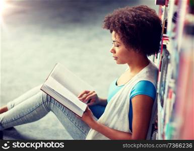 education, high school, university, learning and people concept - african american student girl reading book sitting on floor at library. african student girl reading book at library