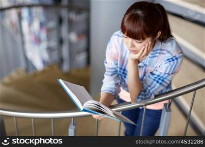 education, high school, university, learning and people concept - asian student girl reading book on stairs at library