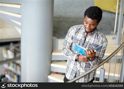 education, high school, university, learning and people concept - happy african american student boy or young man reading book on stairs at library