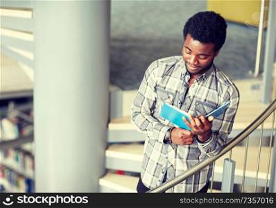 education, high school, university, learning and people concept - happy african american student boy or young man reading book on stairs at library. african student boy or man reading book at library