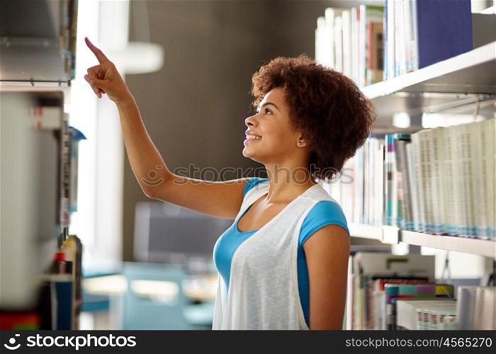education, high school, university, learning and people concept - happy smiling african american student girl pointing finger to book on shelf at library