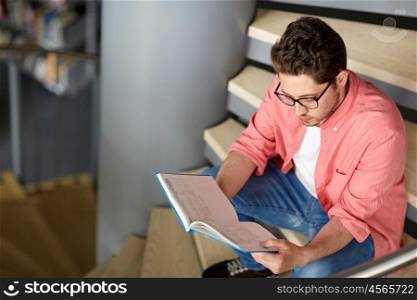 education, high school, university, learning and people concept - student boy or young man reading book sitting on stairs at library