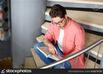 education, high school, university, learning and people concept - student boy or young man reading book sitting on stairs at library