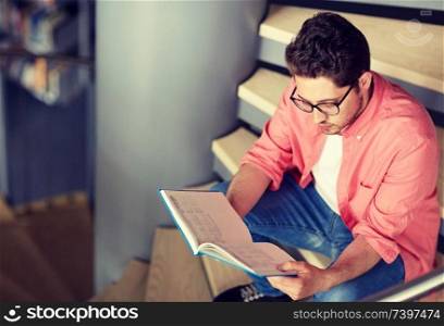 education, high school, university, learning and people concept - student boy or young man reading book sitting on stairs at library. student boy or young man reading book at library
