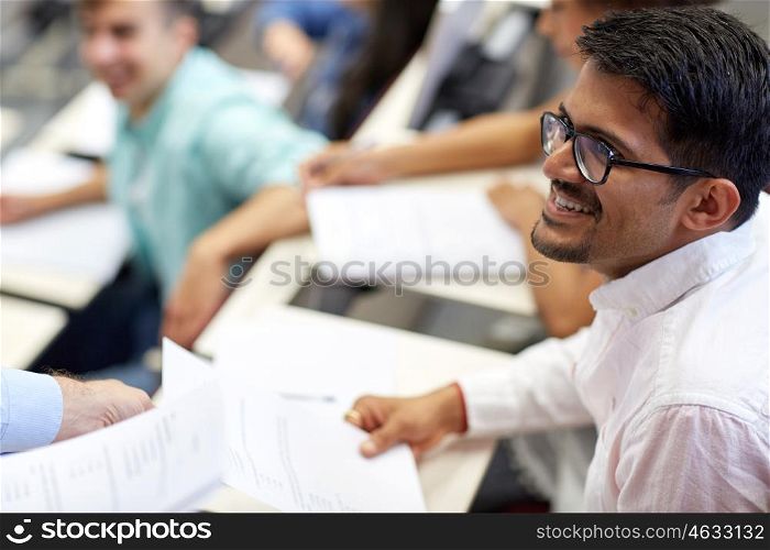education, high school, university, learning and people concept - teacher giving exam test to smiling indian student man at lecture