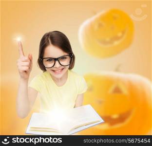 education, holidays, childhood, vision and people concept - smiling little girl in glasses with magic book over halloween pumpkins background