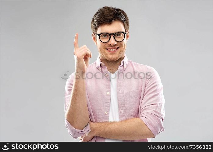 education, idea and people concept - smiling young man in glasses pointing finger up over grey background. smiling young man in glasses pointing finger up