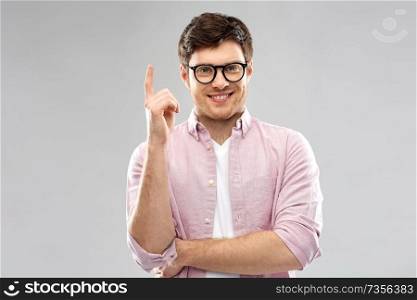 education, idea and people concept - smiling young man in glasses pointing finger up over grey background. smiling young man in glasses pointing finger up