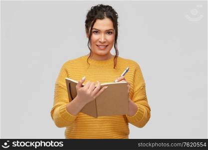 education, inspiration and people concept - happy smiling young woman with pierced nose writing to diary or notebook over grey background. happy young woman writing to diary or notebook