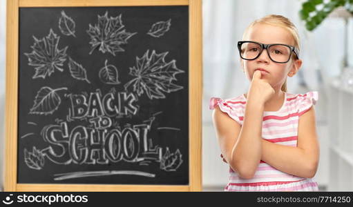 education, learning and people concept - cute little girl in black glasses over chalkboard with back to school lettering on background. little student girl in glasses thinking