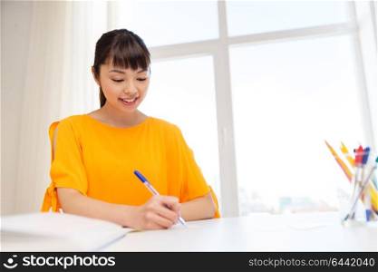 education, learning and people concept - happy asian student girl writing to notebook at home. happy student girl with book and notebook at home