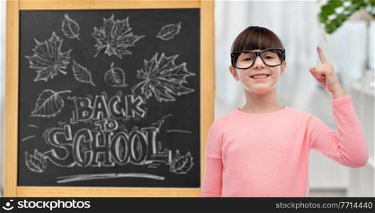 education, learning and people concept - happy little student girl in eyeglasses pointing finger up over chalkboard with back to school lettering on background. little student girl in glasses pointing finger up