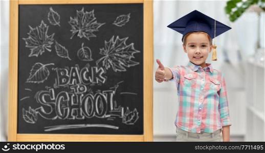education, learning and people concept - happy little student girl with in bachelor hat or mortarboard showing thumbs up over chalkboard with back to school lettering on background. student girl in bachelor hat showing thumbs up