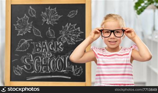 education, learning and people concept - smiling cute little girl in black glasses over chalkboard with back to school lettering on background. little student girl in glasses over chalkboard