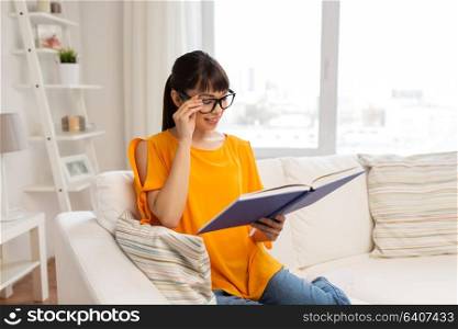 education, literature and people concept - smiling young asian woman in glasses reading book at home. young asian woman in glasses reading book at home