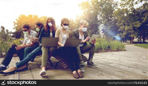 education, pandemic and people concept - group of students with laptop and tablet pc computers wearing face protective medical masks for protection from virus disease at park. teenage students in masks with computers at campus