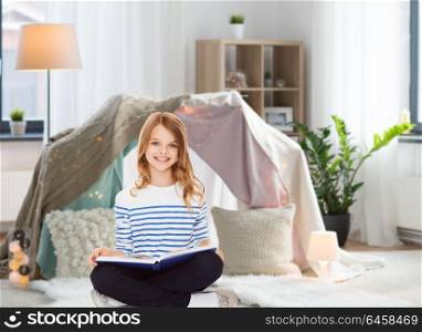 education, people and leisure concept - happy smiling girl reading book at home over kids room and tepee background. happy smiling girl reading book at home