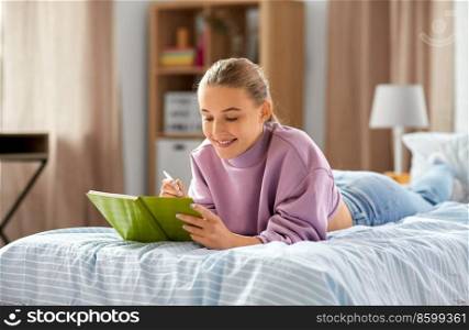education, people and leisure concept - happy smiling student girl with diary and pencil lying on bed at home. girl lying on bed and writing to diary at home