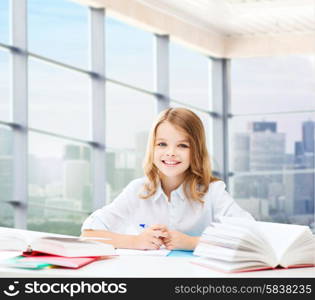 education, people, children and school concept - happy student girl sitting at table with books and writing in notebook over classroom background