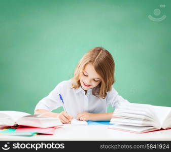 education, people, children and school concept - happy student girl sitting at table with books and writing in notebook over green chalk board background
