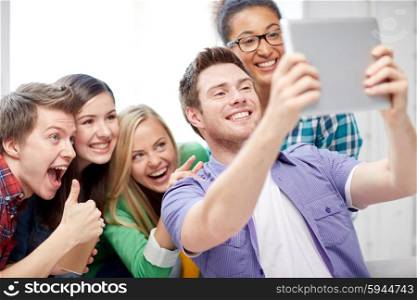 education, people, friendship, technology and learning concept - group of happy international high school students with tablet pc computer taking selfie in classroom and showing thumbs up