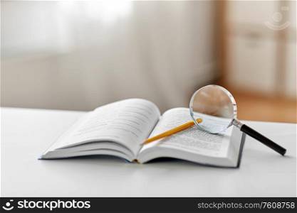 education, school and learning concept - book with magnifier and pencil on table at home. book with magnifier and pencil on table at home