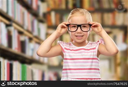 education, school and learning concept - smiling cute little girl in black glasses over library book shelves background. smiling little girl in black glasses at library