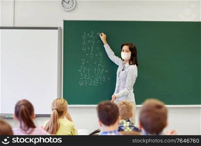 education, school and pandemic concept - female math teacher wearing face protective medical mask for protection from virus disease writing mathematic task on green chalkboard for group of students. teacher in mask writing on chalkboard at school