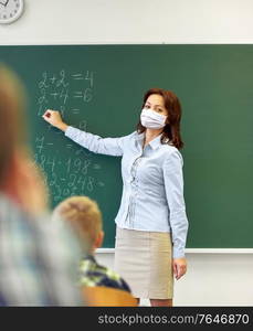 education, school and pandemic concept - female math teacher wearing face protective medical mask for protection from virus disease writing mathematic task on green chalkboard for group of students. teacher in mask writing on chalkboard at school