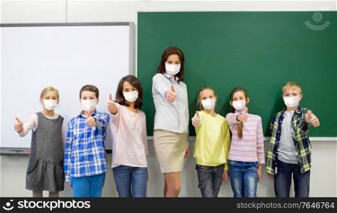 education, school and pandemic concept - group of students and teacher wearing face protective medical mask for protection from virus disease showing thumbs up at classroom. students and school teacher in mask show thumbs up