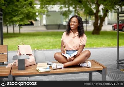education, school and people concept - happy smiling african american student girl with pencil making notes in book in city. happy african student girl making notes in book