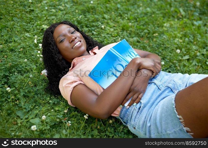 education, school and people concept - happy smiling african american student girl with math textbook lying on grass. african student girl with textbook lying on grass