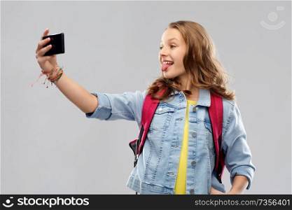 education, school and people concept - happy smiling teenage student girl with bag taking selfie by smartphone and showing her tongue over grey background. teenage student girl taking selfie by smartphone