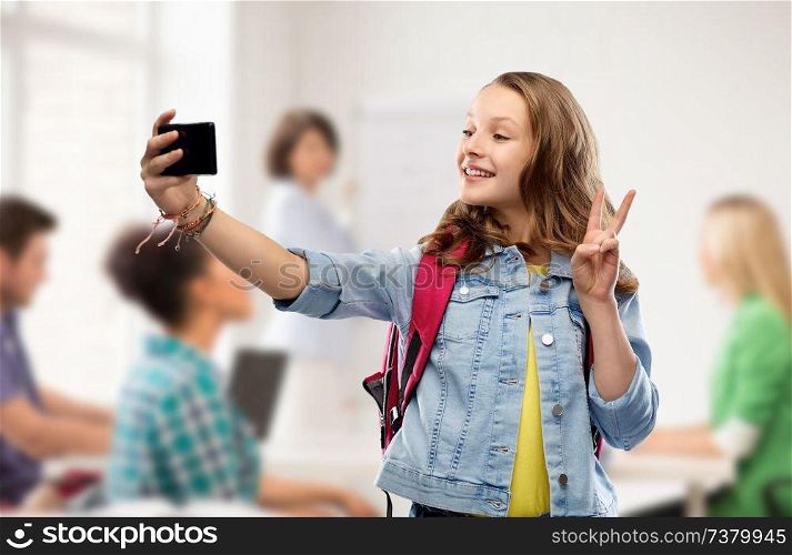 education, school and people concept - happy smiling teenage student girl with bag taking selfie by smartphone and showing peace over classroom background. teenage student girl taking selfie by smartphone