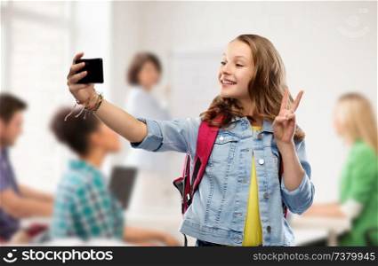 education, school and people concept - happy smiling teenage student girl with bag taking selfie by smartphone and showing peace over classroom background. teenage student girl taking selfie by smartphone