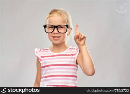education, school and vision concept - smiling cute little girl in black glasses pointing finger up over grey background. smiling little girl in glasses pointing finger up