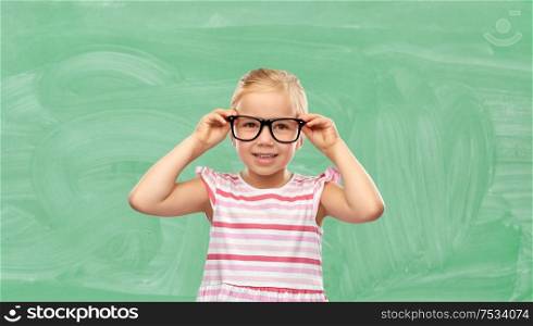 education, school and vision concept - smiling cute little girl in black glasses over green chalkboard background. smiling cute little girl in glasses at school