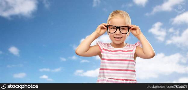 education, school and vision concept - smiling cute little girl in black glasses over blue sky and clouds background. smiling little girl in glasses over sky background