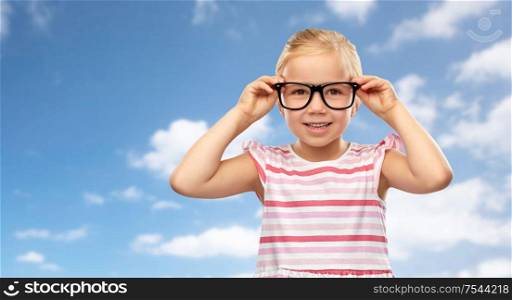 education, school and vision concept - smiling cute little girl in black glasses over blue sky and clouds background. smiling cute little girl in black glasses