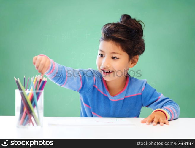 education, school, children, creativity and happy people concept - happy little girl drawing with coloring pencils over green chalk board background