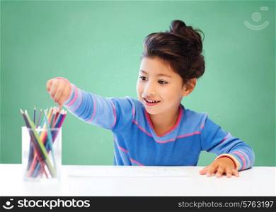 education, school, children, creativity and happy people concept - happy little girl drawing with coloring pencils over green chalk board background