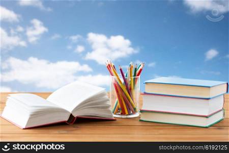 education, school, creativity and object concept - close up of crayons or color pencils and books on wooden table over blue sky and clouds background