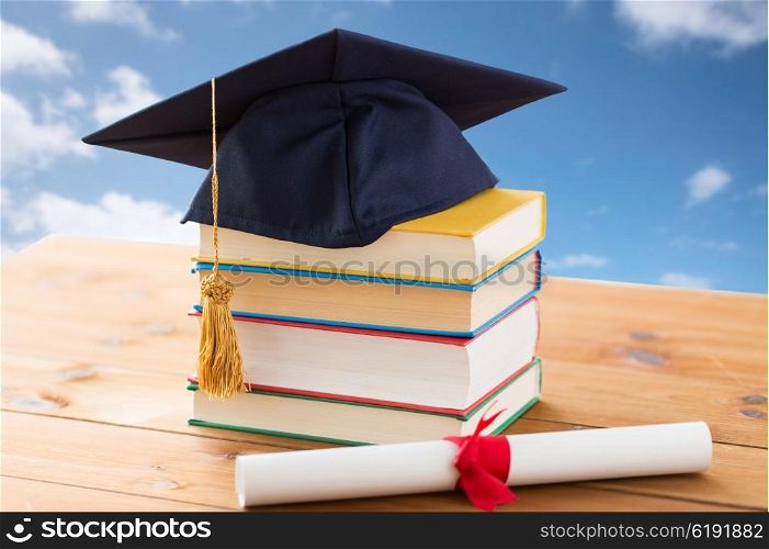 education, school, graduation and knowledge concept - close up of books and mortarboard with diploma on wooden table over blue sky and clouds background