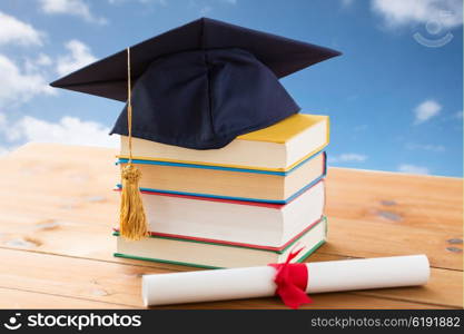 education, school, graduation and knowledge concept - close up of books and mortarboard with diploma on wooden table over blue sky and clouds background